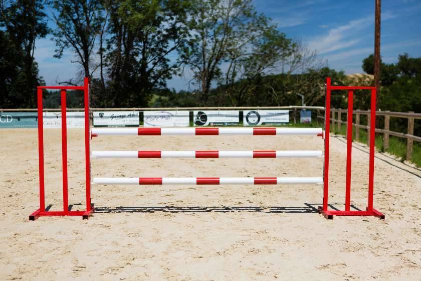 Obstacles d’équitation à allure compétition: barres blanches et rouges montées entre montants rouges sur piste en sable.